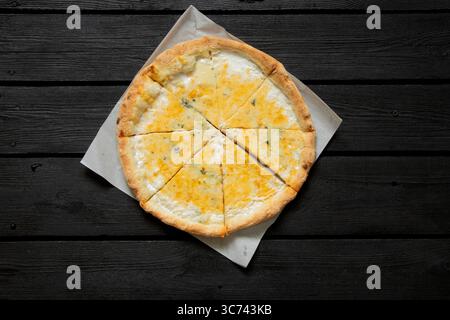 Pizza au fromage entier coupé en morceaux couchés sur du papier sur fond de table en bois noir, vue de dessus, délicieux dîner Banque D'Images