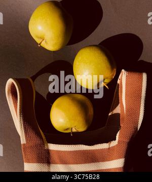Trois pommes vertes et un sac en coton rayé sur une surface blanche avec des ombres douces et une esthétique minimaliste à la lumière naturelle du soleil Banque D'Images
