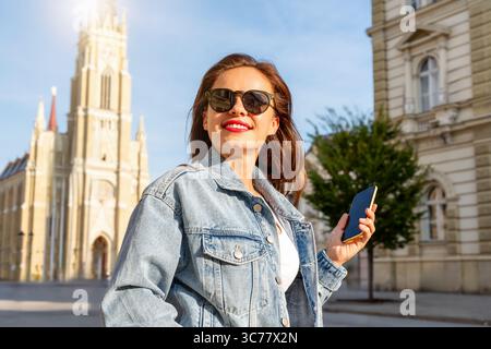 Femme tendance portant une tenue en denim et des lunettes de soleil marchant dans la rue urbaine avec smartphone Banque D'Images