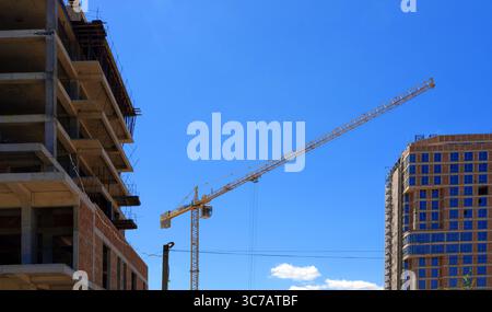 Haute grue à tour jaune opérant entre un bâtiment inachevé et une structure moderne en construction contre un ciel bleu clair. Banque D'Images