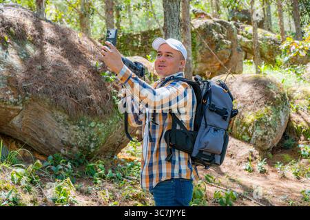 Photographe colombien joyeux prend un selfie dans la forêt tout en faisant face à l'appareil photo, tenant son reflex numérique et portant un grand sac à dos, profitant d'un ensoleillement Banque D'Images