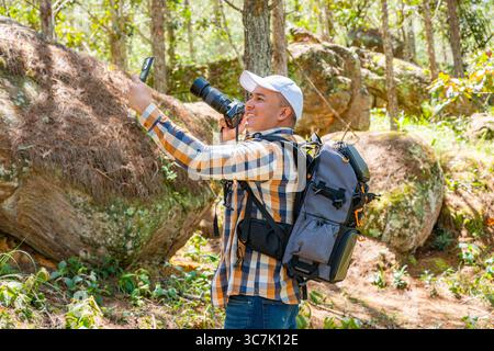 Photographe latino-américain prend un selfie latéral dans une forêt de pins. Il sourit en toute confiance, tenant son appareil photo reflex numérique dans une main tout en portant un dos Banque D'Images