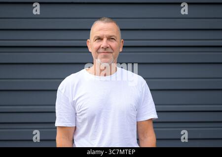 Homme mature confiant avec les cheveux gris courts et la barbe, debout devant un mur gris foncé moderne. Banque D'Images