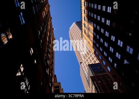 Une flèche ensoleillée se dresse entre des façades ombragées de briques et de verre dans un étroit canyon de Manhattan, capturant le rythme vertical dynamique de la ville. Banque D'Images