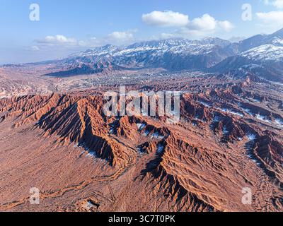 Un drone aérien capture le paysage panoramique des collines rouges et des ravins de mars Canyon, menant l'œil vers une ville lointaine et des montagnes enneigées Banque D'Images