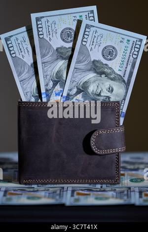 Portefeuille en cuir éclairé latéralement plein de monnaie américaine sur une table couverte de billets de cent dollars Banque D'Images