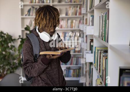 Étudiant masculin concentré avec livre de lecture de cheveux afro près de l'étagère Banque D'Images