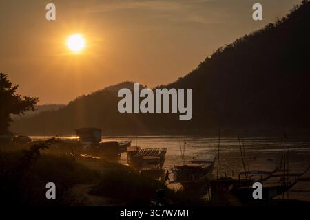 Coucher de soleil doré sur des bateaux traditionnels amarrés sur le fleuve Mékong avec des silhouettes de collines en arrière-plan, Laos. Banque D'Images