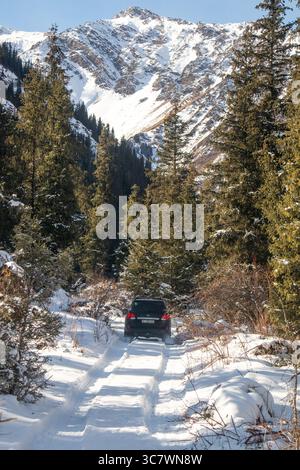 Naryn, Kirghizistan - 12 octobre 2024 : un véhicule hors route sombre roule sur une route enneigée à travers une forêt avec une montagne enneigée en arrière-plan Banque D'Images