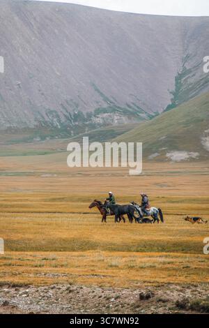 Naryn, Kirghizistan - 12 octobre 2024 : deux hommes kirghizes à cheval et un chien marchent sur une vaste prairie dorée et verte avec une grande montagne accidentée dans le Banque D'Images