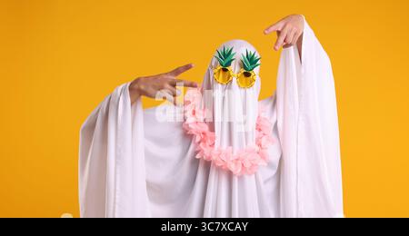 Fantôme drôle. Homme couvert de feuille blanche dans des lunettes de soleil et couronne de fleurs gestuelles sur fond orange Banque D'Images