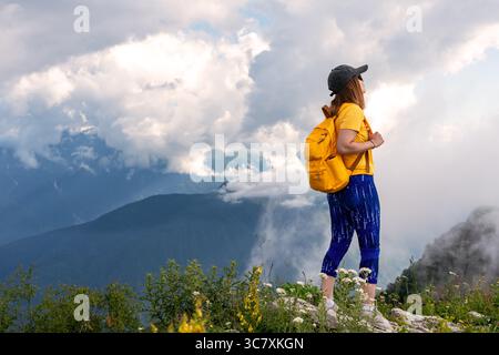 Jeune femme randonneur en chemise jaune et sac à dos se dresse sur le pic de montagne rocheuse, regardant un paysage à couper le souffle avec des nuages et des montagnes lointaines, embo Banque D'Images