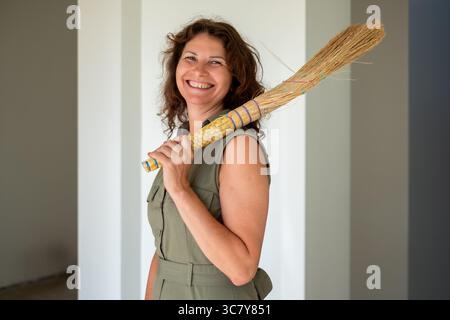 Femme joyeuse aux cheveux bouclés tient un balai à l'intérieur moderne, portant une robe verte, rayonnant positivité et chaleur dans un environnement lumineux Banque D'Images