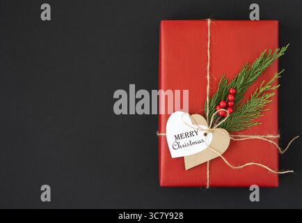 Boîte d'artisanat rouge avec étiquette cadeau en forme de coeur avec texte joyeux noël, brindille de cèdre et baies sur un fond sombre. Espace pour le texte. Banque D'Images