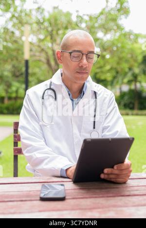 Docteur masculin chauve en uniforme médical blanc avec stéthoscope assis à l'extérieur dans le jardin de l'hôpital. Souriant, confiant et détendu. Concept de soins de santé Banque D'Images