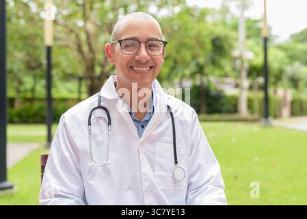 Docteur masculin chauve en uniforme médical blanc avec stéthoscope assis à l'extérieur dans le jardin de l'hôpital. Souriant, confiant et détendu. Concept de soins de santé Banque D'Images