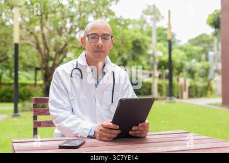 Docteur masculin chauve en uniforme médical blanc avec stéthoscope assis à l'extérieur dans le jardin de l'hôpital. Souriant, confiant et détendu. Concept de soins de santé Banque D'Images
