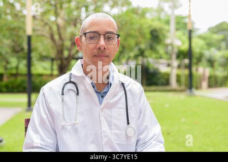 Docteur masculin chauve en uniforme médical blanc avec stéthoscope assis à l'extérieur dans le jardin de l'hôpital. Souriant, confiant et détendu. Concept de soins de santé Banque D'Images