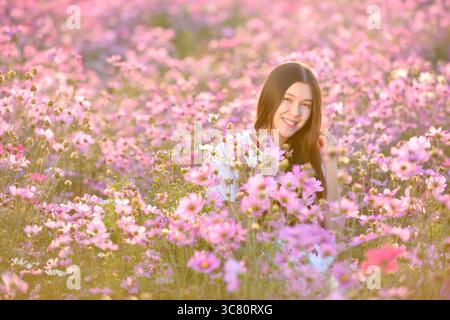 Portrait d'une jeune femme souriante assise dans un champ de fleurs roses du cosmos (Cosmos bipinnatus), Thaïlande Banque D'Images