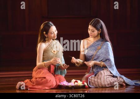 Deux jeunes femmes portant des vêtements traditionnels thaïlandais assis côte à côte sur le sol faisant des guirlandes florales (Phuang Malai) Banque D'Images