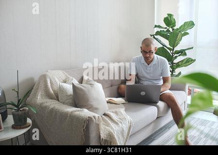 Homme travaillant à la maison, en utilisant un ordinateur portable sur un canapé dans un salon confortable Banque D'Images
