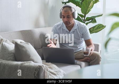 Homme travaillant à domicile ayant un appel vidéo avec un ordinateur portable et un casque tout en buvant du café Banque D'Images