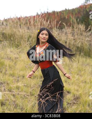 Jeune femme adulte se déplaçant gracieusement à travers un champ herbeux dans une robe fleurie brodée vibrante, avec de longs cheveux coulant dans la brise et une fleur dedans Banque D'Images