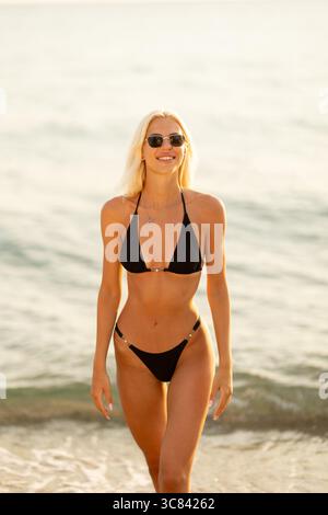 Une femme joyeuse se promène le long de la plage de sable, vêtue d'un élégant bikini et de lunettes de soleil. Les vagues douces reflètent les couleurs chaudes du coucher de soleil, cr Banque D'Images