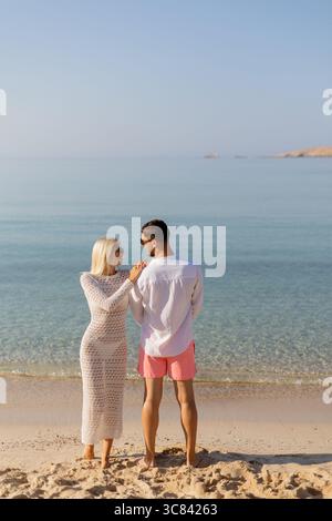 Un couple se tient près de la plage, profitant des eaux calmes et de belles teintes de coucher de soleil. Leur attitude détendue montre l'intimité comme ils partagent un Banque D'Images