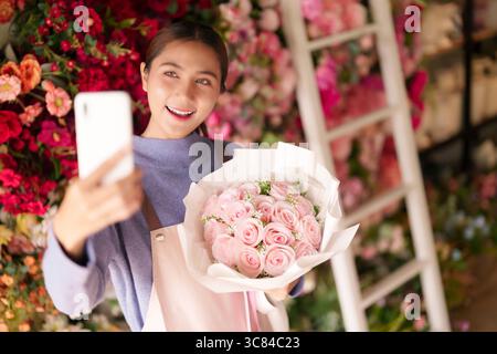 Jeune femme asiatique tient joyeusement bouquet de rose rose fraîche prenant selfie avec smartphone à l'intérieur d'un magasin de fleurs vibrant favorisant les affaires Banque D'Images