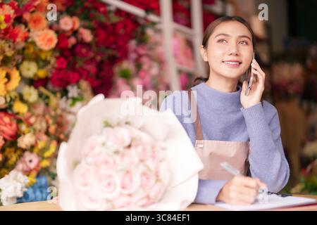 Fleuriste jeune adulte asiatique dans un magasin de fleurs vibrant. Gestion des appels des clients, rédaction de notes pour les commandes de fleurs personnalisées. Dédié au service. Banque D'Images