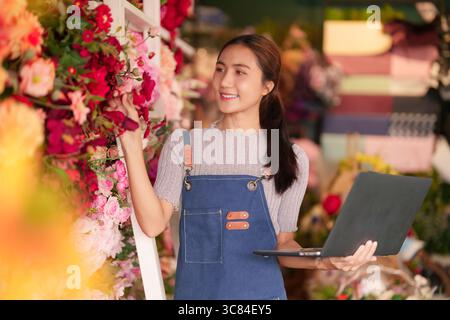 Femme asiatique adulte fleuriste gère efficacement son beau magasin de fleurs à l'aide d'un ordinateur portable. Elle sourit tout en arrangeant des fleurs fraîches colorées. Banque D'Images