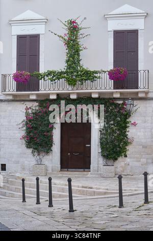 Charmante entrée de villa européenne avec fleurs de bougainvilliers violets, porte en bois sombre, balcon avec volets et architecture en pierre Banque D'Images