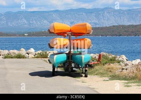 Six kayaks - quatre orange et deux turquoises - sont empilés sur un porte-remorque sur un chemin pavé au bord du lac, avec un rivage rocheux et une chaîne de montagnes lointaine Banque D'Images