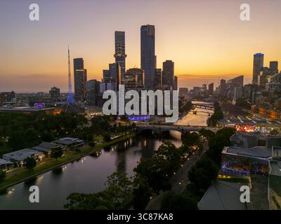 Vue aérienne des gratte-ciel dynamiques de la ville qui se reflète dans la rivière Yarra, avec la flèche du Centre des arts et Federation Square qui se démarquent face au chaud coucher de soleil, Melbourne, Victoria, Australie. Banque D'Images