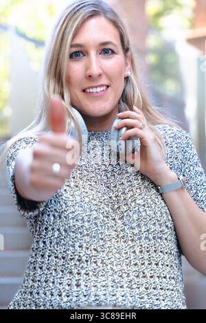 Femme heureuse donnant les pouces vers le haut, tenant le smartphone, geste positif, souriant à l'extérieur Banque D'Images