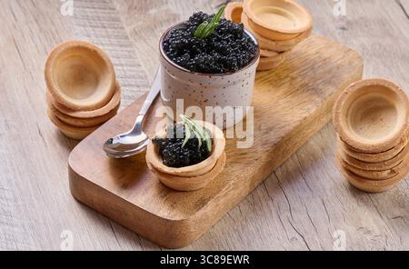 Caviar noir, dans un petit pot en céramique et mini tartelette, placé sur une planche de bois. La composition comprend une cuillère et des piles de tartelettes vides Banque D'Images