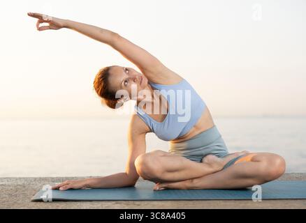 Heureuse jeune femme expérimentée professeur de fitness effectue le yoga parshvottanasana assis sur la plage le matin Banque D'Images