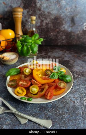 Vue de face de tomates mélangées, basilic frais, bouteille d'huile d'olive et moulin à poivre en bois sur fond brun bleu foncé Banque D'Images