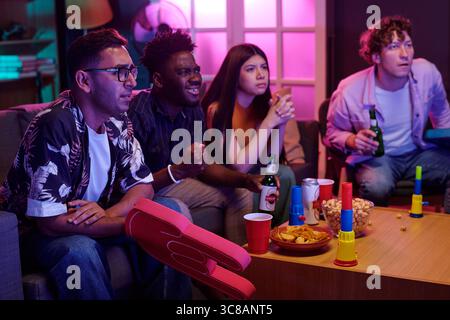 Groupe de jeunes amis multiethniques adultes regardant la télévision ensemble, assis sur le canapé, tenant des boissons et des collations, concentré sur l'écran, s'engageant dans une activité sociale à l'intérieur Banque D'Images