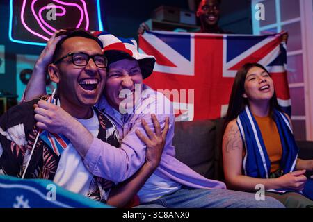 Groupe multiethnique de jeunes adultes célébrant tout en regardant un événement sportif à la télévision, homme caucasien portant un chapeau embrassant un homme sud-asiatique, femme asiatique souriante, drapeau britannique en arrière-plan Banque D'Images