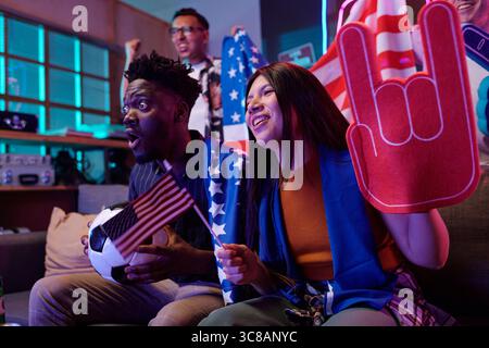 Jeune homme noir tenant un ballon de football et un petit drapeau américain assis à côté de la jeune femme asiatique acclamant avec la main en mousse tout en regardant un événement sportif, homme caucasien d'âge moyen excité debout derrière Banque D'Images