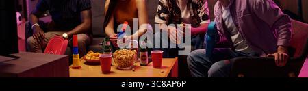 Groupe de jeunes adultes et adolescents assis sur le canapé tenant des boissons et des souffleurs de fête, regardant la télévision, partageant des collations et du pop-corn, des amis multiethniques appréciant de se rassembler à l'intérieur Banque D'Images