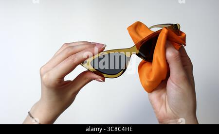Le processus de nettoyage des lunettes de soleil. Les mains féminines tiennent des lunettes triangulaires. Une éponge orange essuie le verre. Verre noir. Cadre marron translucide. Blanc t Banque D'Images