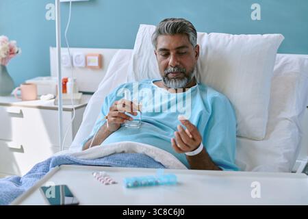 Homme biracial d'âge moyen couché dans un lit d'hôpital prenant des médicaments avec de l'eau Banque D'Images