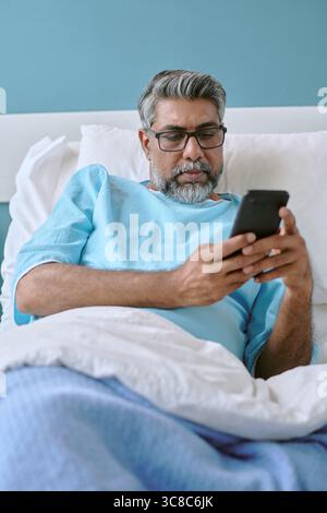 Homme biracial d'âge moyen couché dans le lit d'hôpital à l'aide d'un smartphone Banque D'Images