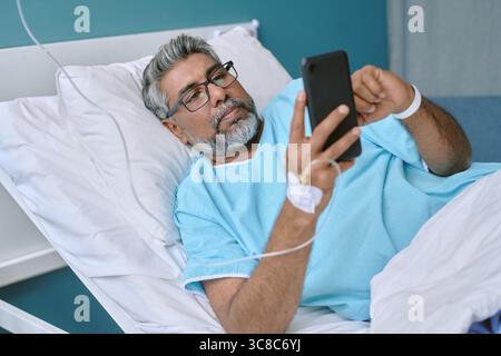 Homme biracial d'âge moyen couché dans le lit d'hôpital à l'aide d'un appareil smartphone Banque D'Images