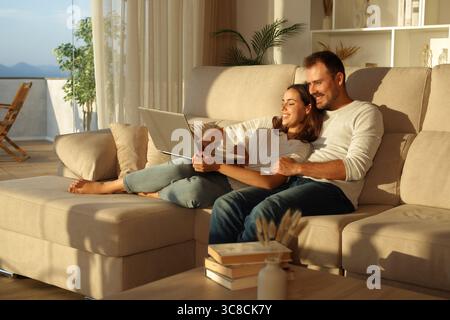 Couple heureux à la maison en utilisant un ordinateur portable ensemble assis sur un canapé au coucher du soleil Banque D'Images