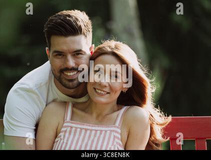 Heureux jeune couple dans un parc. Homme embrassant une belle femme par derrière alors qu'elle est assise sur un banc rouge Banque D'Images