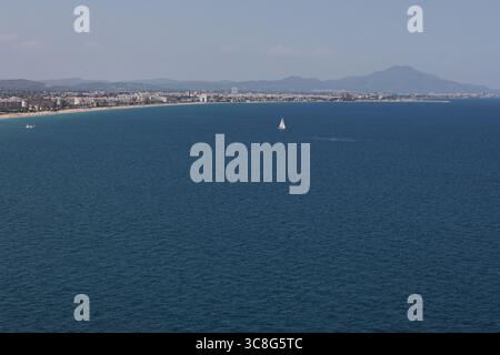 Une large vue panoramique sur la côte montre un voilier glissant à travers la mer bleue calme, avec une ville de plage tentaculaire et des montagnes lointaines sous un cl Banque D'Images